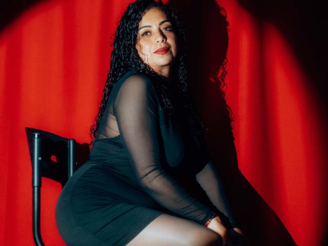 Curvy AlissaaMilf with long black curly hair, wearing a sheer black dress, sitting on a black chair, red curtain background, shadow on her right, smiling, red lipstick.
