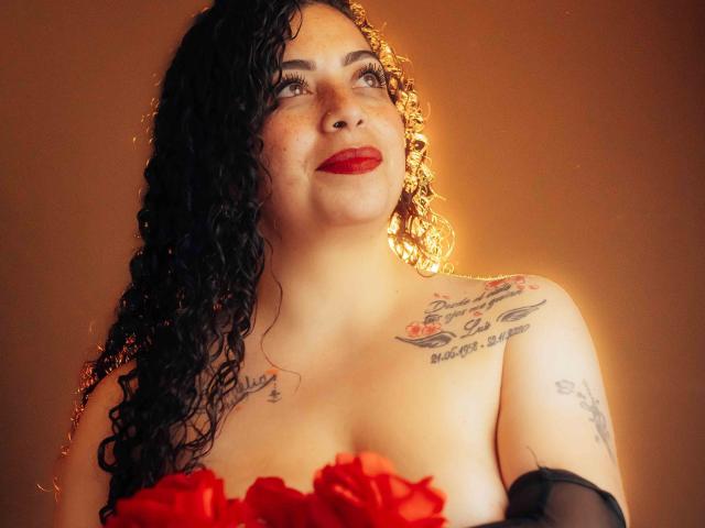 AlissaaMilf with curly black hair, red lipstick, and tattoos, holding red roses, has fair skin and a confident smile, with soft light highlighting her upper body.