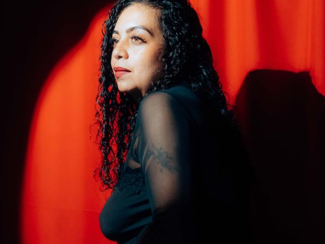 AlissaaMilf with curly black hair, wearing a black sheer top, has a tattoo on her arm, and is looking to the side with a serious expression, in bright red light.
