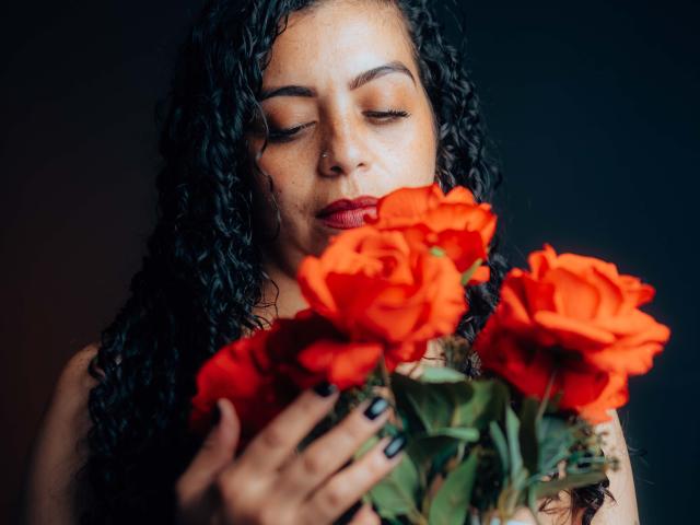 AlissaaMilf with curly black hair, nose ring, and red lipstick, holding bright red roses, eyes closed, black nail polish, dark background, soft lighting, gentle expression.