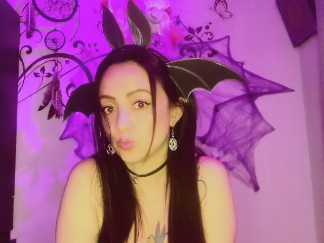 CatalinaMonyque with black bat ears, long black hair, pouty lips, choker, earrings, and bat wings, in purple-lit photo, looking at camera, showing off her tits.