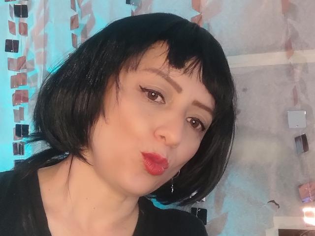 CatalinaMonyque, com cabelo preto curto, batom vermelho e delineador, fazendo beicinho com seus lábios cheios, vestindo uma camisa preta, com um rosto bonito, sexy e atraente..