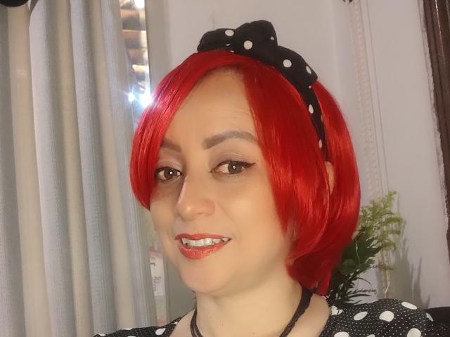 CatalinaMonyque with bright red bob, wearing black polka dot headband, smiling with white teeth, brown eyes, and black polka dot top, has light skin and subtle makeup.