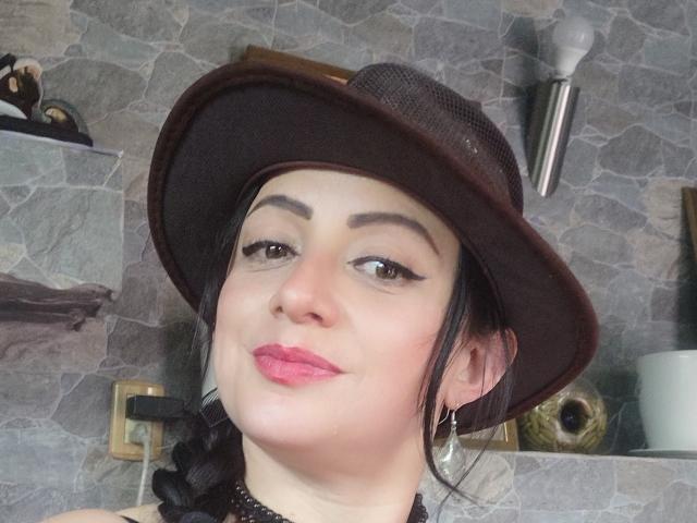 CatalinaMonyque, with a brown hat, pink lipstick, and black braid, smiling at the camera, showing her tits slightly with a choker and hoop earrings.