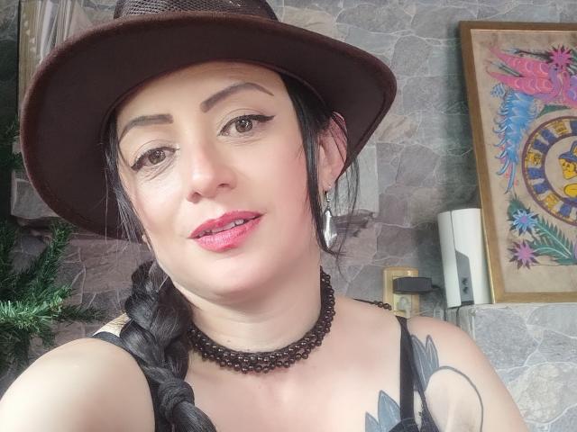 CatalinaMonyque mit dunklem, geflochtenem Haar, einem braunen Hut, pinkem Lippenstift, einer schwarzen Halskette, einem Tattoo an der rechten Schulter, Creolen und einem Lächeln, die in einem Selfie ihre Brüste zeigt..