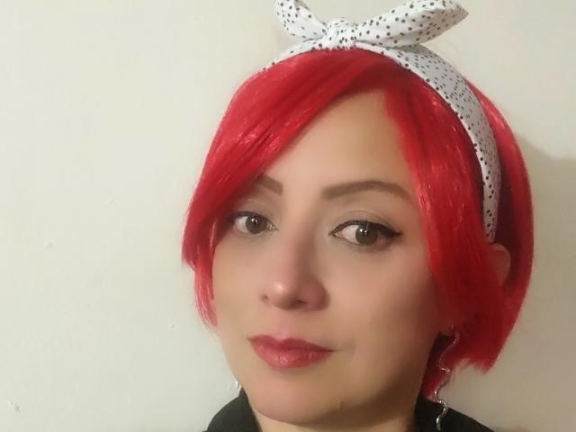 CatalinaMonyque has bright red hair, a white polka-dot headband, and is wearing red lipstick. Her brown eyes are slightly upturned. She's looking at the camera with a small smile.