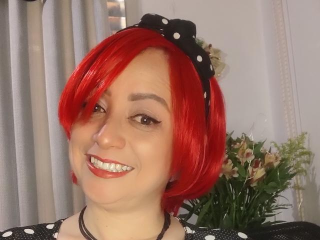 CatalinaMonyque with bright red bob, wearing a black polka dot headband, smiling with red lipstick, showing her teeth, wearing a black and white polka dot top.