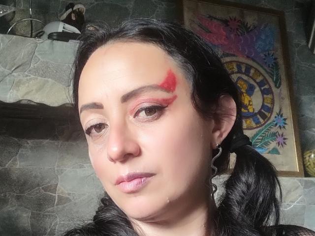 CatalinaMonyque has black hair in pigtails, red eyeshadow with a matching red triangle on her forehead, and pink lipstick. Her skin is light, and she's wearing a black top.