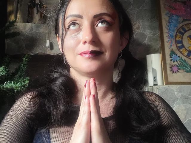 CatalinaMonyque with dark hair in pigtails, wearing sheer black top, hands pressed together, red lipstick, looking up, shiny skin, red nail polish, stone wall background, decorative clock.