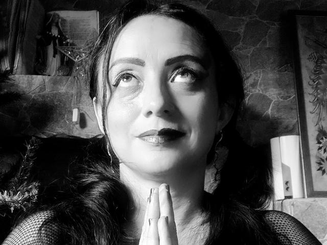 Black-and-white photo of CatalinaMonyque with dark hair, hoop earrings, and glossy lipstick, looking up with a slight smile, hands together, wearing a fishnet top.