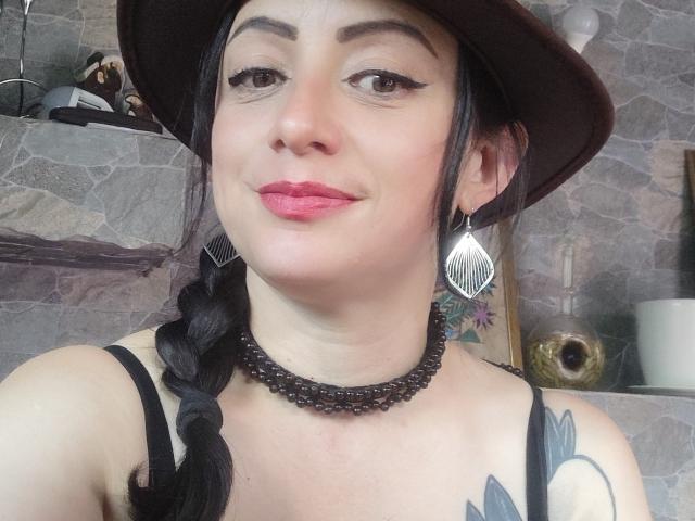 CatalinaMonyque with black braided hair, brown hat, red lipstick, black eyeliner, silver earrings, black beaded necklace, and tattoo on right shoulder, smiling slightly. Boobs visible in black top.