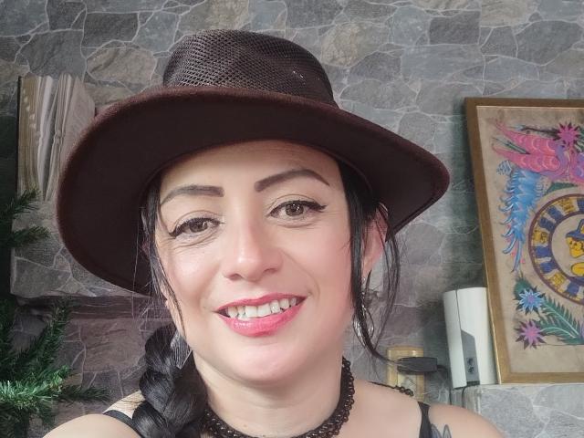 CatalinaMonyque with dark brown hat, black braided hair, pink lipstick, and black necklace, smiling at camera, stone wall in background, floral painting on right.