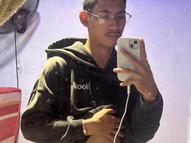 HiddenDesireAru taking a selfie in a mirror, wearing glasses and a dark hoodie, holding a white iPhone, with a fan and red-and-white chair in the background.