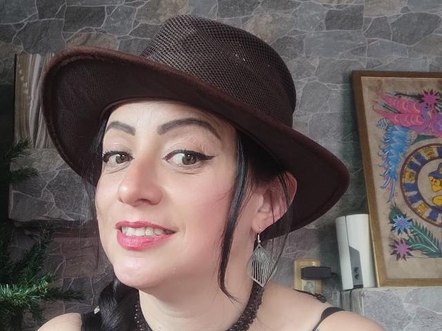 CatalinaMonyque with brown mesh hat, black braided hair, pink lipstick, and eyeliner, smiling at camera, stone wall background, colorful framed art on right.