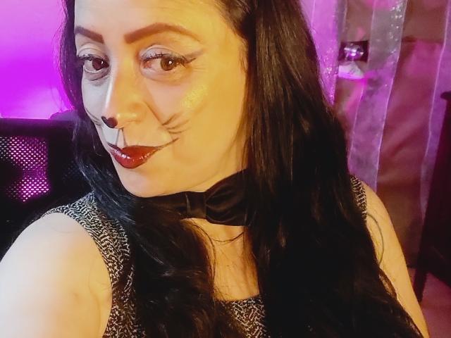 CatalinaMonyque with long black hair, dark lipstick, black nose ring, and black collar, wearing a patterned dress, smiling slightly, in a purple-lit room.