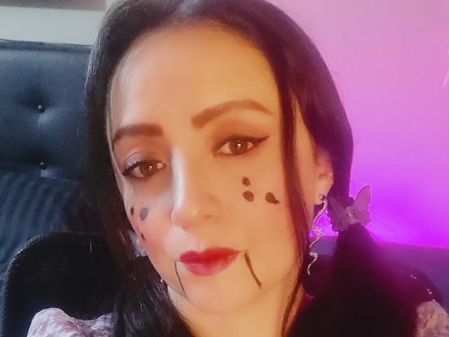 CatalinaMonyque with black makeup dots on her cheeks, red lipstick, and butterfly earrings, has brown eyes and dark brown hair, looking at the camera with a slight smile.