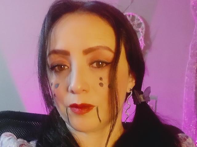 CatalinaMonyque with dark hair, red lipstick, black face paint dots, brown eyes, black eyebrows, butterfly earrings, and pigtails, sitting in a chair with a pinkish-purple light.