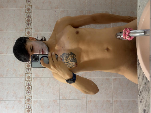Dallas, nue, tenant un téléphone pour un selfie dans un miroir, tatouage visible sur la poitrine, godemichet rose dans la main droite, portant un bracelet noir, cheveux noirs courts, peau claire, petit pénis, carrelage de salle de bain..