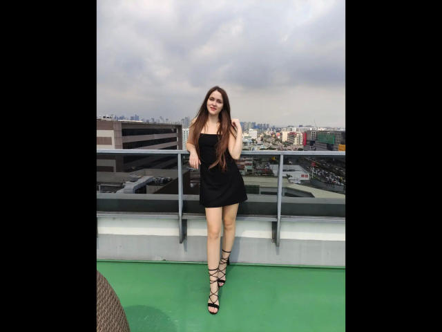 KnackigeOssi-hot standing on a rooftop, wearing a black strapless mini-dress and black lace-up sandals, with long brown hair, smiling, and cloudy sky in the background.
