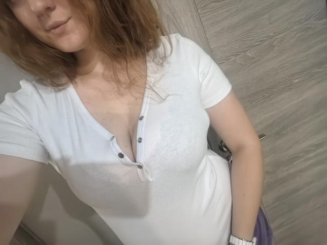 ArianaXsmile, with wavy red hair, wearing a white ribbed, button-down, short-sleeve shirt, showing her tits and a white wristwatch, in a slightly angled selfie.