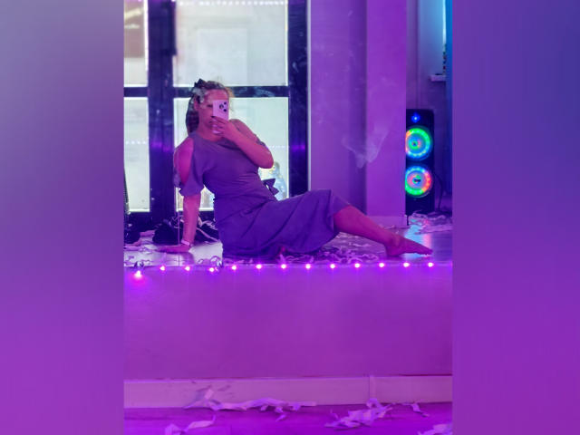 ArianaXsmile is sitting on a purple-lit windowsill in a purple dress, smoking, with her tits partially visible, holding a phone, and surrounded by white petals.