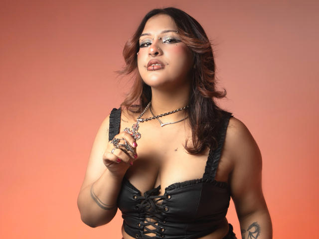 DanniLuke has medium brown skin, wavy dark red hair, and wears a black lace-up top, showing her boobs. She's holding a ring, smiling, with pink nail polish.