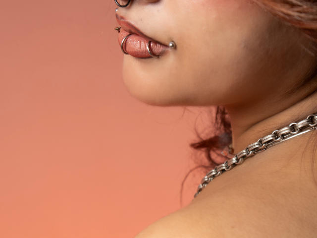DanniLuke's face with a metal nose ring, lip piercing, and chain necklace, showing a side profile with slightly parted pinkish lips. Her skin is light and she has red hair.