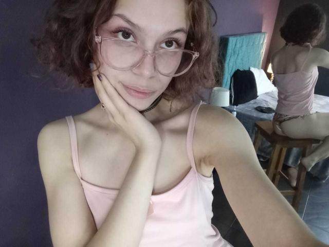 SophieBlake with wavy brown hair, pink glasses, pink tank top, choker, and hand on cheek, showing her tits in a mirror selfie, pink nipples visible.