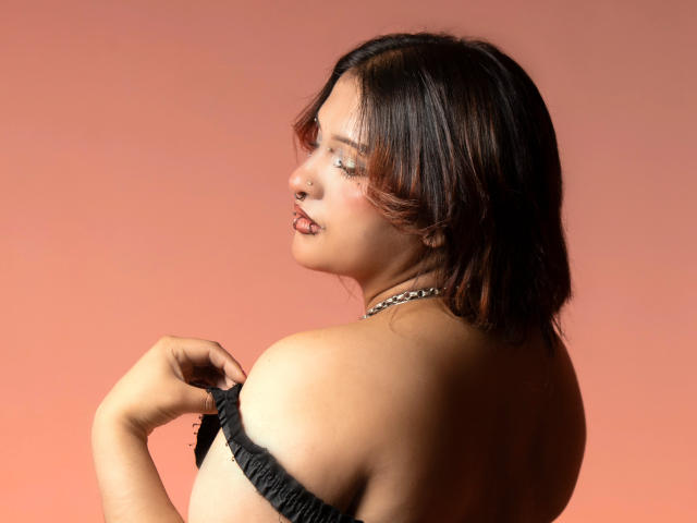 DanniLuke with short brown hair, nose ring, and sparkly eyeshadow, pulling down a black strap, showing her bare shoulder and upper breast. She's wearing a chain necklace.