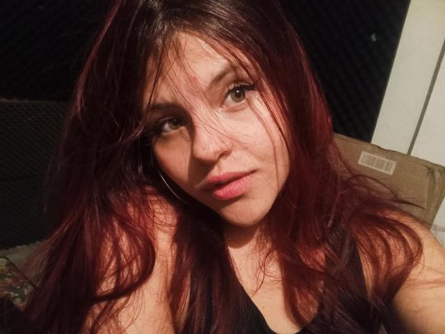 SophieHill with red hair, looking into the camera, has a serious expression, wearing a black top, showing her bare shoulders and a slight tan.