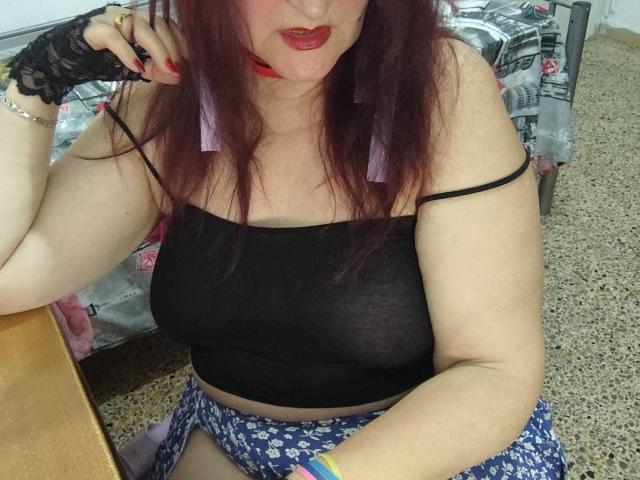 Sabrhina with red lipstick, black sheer top showing her tits, purple earrings, lace finger, floral skirt, sitting on carpet, holding phone to ear, looking at camera.