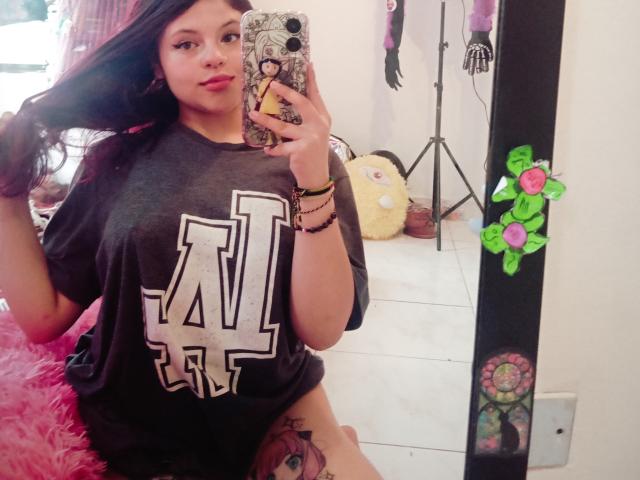 SophieHill with long dark hair, wearing a black "U" t-shirt, holding a phone with a patterned case, showing a thigh tattoo, and pink fluffy chair in background.