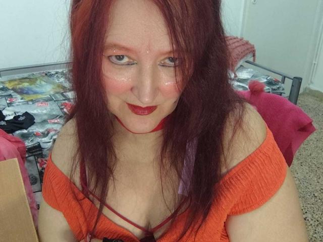 Sabrhina with red hair, wearing an orange off-shoulder top, red lipstick, and a red choker, smiling at the camera, showing her tits and a cute ass.