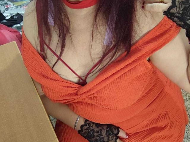 Sabrhina with purple hair, red choker, and orange dress showing cleavage with red straps, wearing black lace glove, holding her chest. Her tits are visible under the dress.
