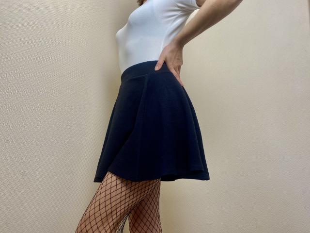 Rorria veste uma blusa branca, uma saia preta e meias de rede, e está em pé com uma mão na cintura, exibindo suas nádegas. Sua pele é clara..