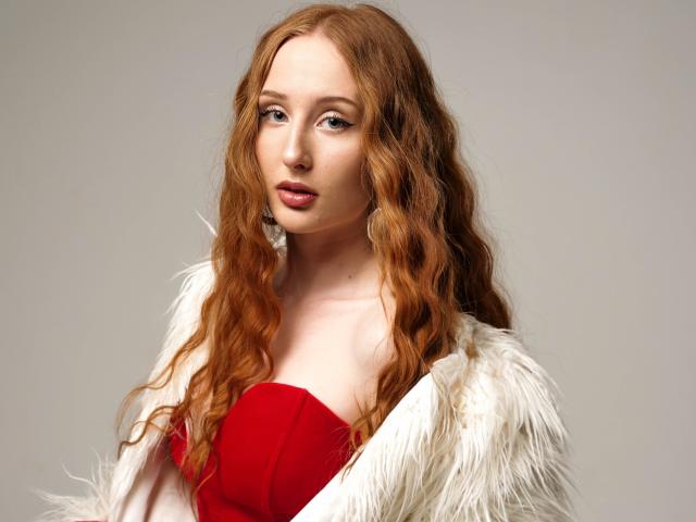TinaSerenade with long red wavy hair, wearing a white fur coat over a red strapless dress, has pink lipstick and a slightly parted mouth, looking directly at camera.