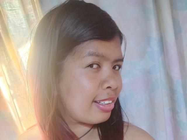MariahSquirting69, femme asiatique aux cheveux bruns foncés, lèvres légèrement entrouvertes laissant apparaître les dents, peau claire, petit nez, yeux marrons, sourire discret, lumière du soleil venant de la gauche, fond pastel doux..