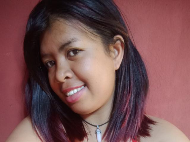MariahSquirting69 met donkerbruin haar, tot op de schouders, met roze highlights, die lichtjes glimlacht, draagt een zwarte choker met een klein hangertje, op een rode achtergrond..