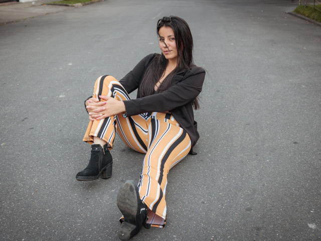 MargarettCooper is sitting on a gray asphalt road, wearing striped orange and black pants, black boots, and a black jacket, with her hands on her knees, looking serious.