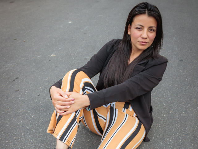 MargarettCooper with long black hair, sitting on gray pavement, wearing a black jacket and yellow-striped pants, hands on knees, looking slightly to the side with a neutral expression.