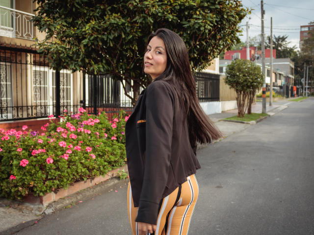 MargarettCooper stands on a street, looking over her shoulder, with long dark hair, black jacket, and yellow-striped pants, showing her ass, smiling. Pink flowers and black fence.