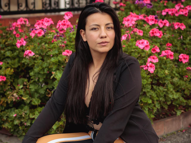 MargarettCooper with long black hair, brown eyes, and medium tits, wearing a black jacket, sitting in front of pink flowers, looking slightly to the side with a neutral expression.