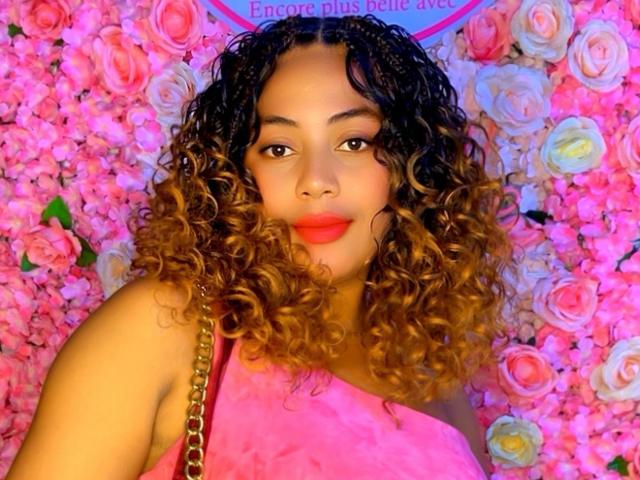 FameSweet has curly brown hair, red lipstick, and is wearing a pink top with a gold chain. Pink roses fill the background. She looks cute and sexy.