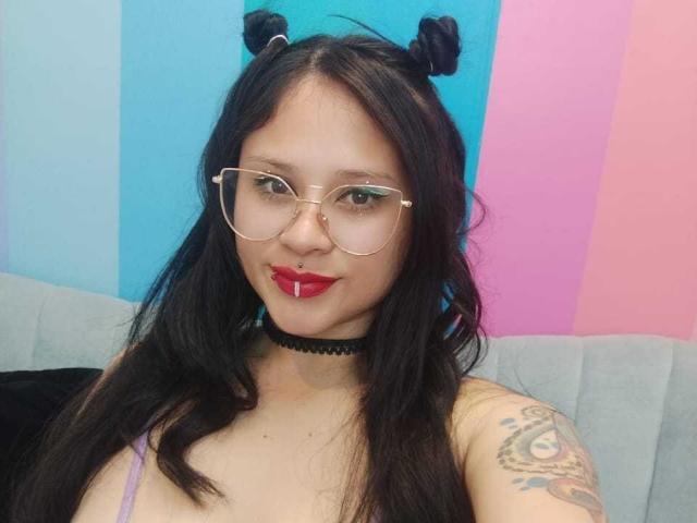 MaraVixen has dark hair in double buns, wearing glasses, a choker, and a purple top, with red lipstick and a nose ring, showing a tattoo on her right arm.