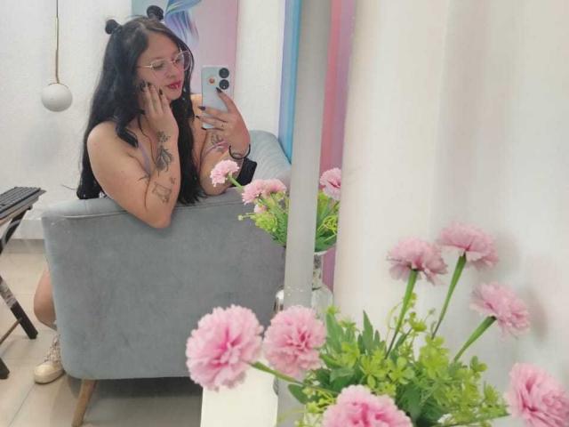 MaraVixen with long black hair, glasses, and cat ears, taking a selfie in a mirror, sitting on a gray chair, with pink flowers in the foreground, and tattoos.