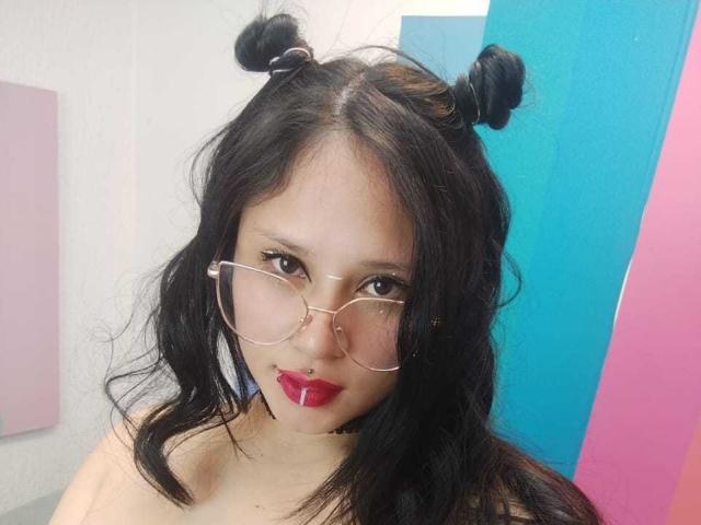 MaraVixen with black hair in double buns, wearing clear glasses and red lipstick, has a nose ring and a black choker, showing her tits.
