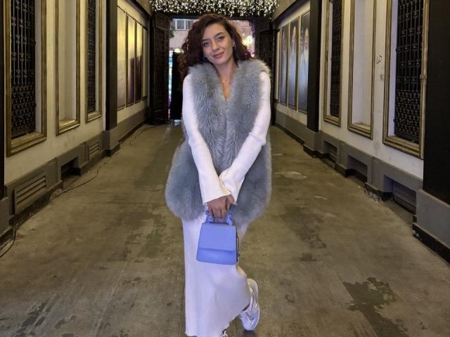 BubbleHeat with curly brown hair, white dress, grey furry vest, holding blue purse, standing in hallway, smiling, wearing white sneakers, concrete floor, ornate walls.