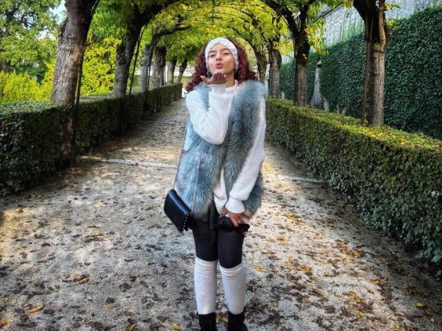 BubbleHeat with brown wavy hair, wearing a white knit sweater, gray fur vest, black pants, white knee socks, and black boots, holding a black clutch, pouting in a park.