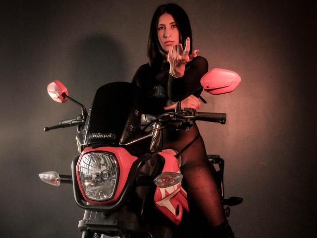 BondageGirlKs with black hair, wearing a black mesh top and sheer black panties, is on a red and black motorcycle, flipping a finger, with a serious expression.