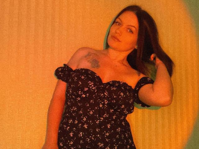 AnastasiaMorante with medium tits, wearing a black floral dress, has a tattoo on her right shoulder, brown hair, and a hand in her hair, looking slightly to the side.
